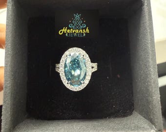 May include: An oval-cut, light blue gemstone ring with a halo of smaller, clear stones. The ring is set in a silver band and presented in a black jewelry box. The brand logo "Hetvansh Jewel" is visible.