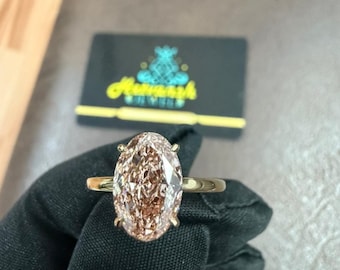 May include: A gold ring featuring a large, oval-cut, light brown gemstone. The ring is held by a gloved hand, with a business card in the background that has the text "Amanish Jewels".