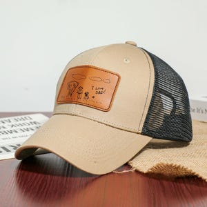 Op de afbeelding: Beige baseballpet met een zwarte mesh achterkant. De voorkant heeft een bruin leren patch met een familietekening en de tekst "I Love DAD!" in zwart. De pet heeft een gebogen klep en een knoop aan de bovenkant.