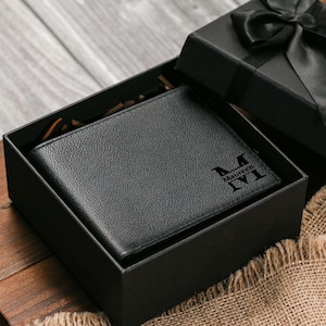 May include: A black leather wallet with a personalized monogram and name is presented in a black gift box. The box is open, revealing the wallet, and is tied with a black satin ribbon. The wallet is on a wooden surface.