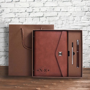 Puede incluir: Un juego de diario y bolígrafo de cuero marrón en una caja de regalo. El diario tiene un cierre de solapa con un cierre plateado y un monograma en forma de diamante. El juego incluye un bolígrafo y una recarga.