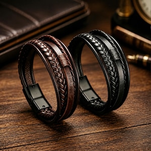 Pode incluir: Duas pulseiras de couro de várias camadas, uma em castanho escuro e outra em preto, cada uma com um cordão de couro entrançado, uma tira de couro liso e um fecho magnético de metal preto. A pulseira castanha tem a data 03.09.2025 gravada no couro.