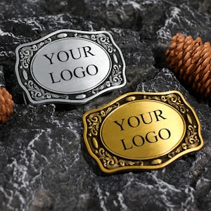 Peut inclure: Deux boucles de ceinture rectangulaires aux coins arrondis et aux bordures ornées de volutes. L'une est argentée avec une finition brossée, l'autre est dorée avec une finition lisse. Les deux portent le texte noir YOUR LOGO au centre.