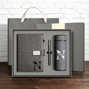 May include: A gray gift set featuring a notebook, pen, and tumbler, all with the name "Nicole" printed on them. The notebook has a magnetic closure. The tumbler is cylindrical. The set is presented in a gray gift box with a matching gift bag.