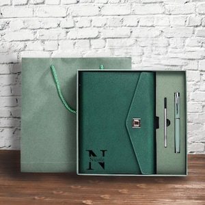 May include: A green journal and pen set in a gift box. The journal has a flap closure and the name "Nicole" printed on the front. The set includes a pen and a refill. The gift bag is green with a green rope handle.