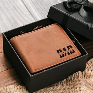 May include: A brown leather wallet with the word "DAD" and two hearts engraved on the front, presented in a black gift box with a black ribbon. The wallet is a bi-fold style.