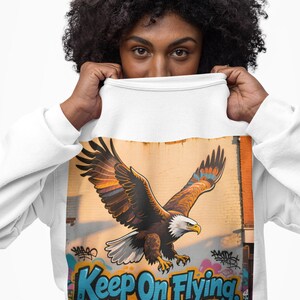 May include: White sweatshirt with a graphic of a flying eagle, set against a graffiti-covered brick wall. The eagle has brown, orange, and blue wings. The text "Keep On Flying, Don't Look Down" is written in blue and white.