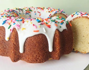 May include: A golden-brown bundt cake with white icing and colorful sprinkles. A slice has been cut, revealing the cake's interior. The cake sits on a white surface, ready to be served.