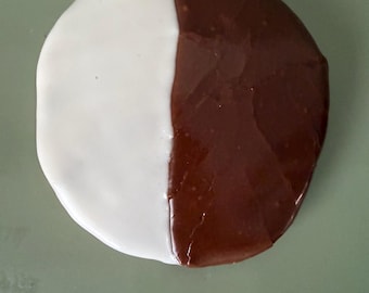 Puede incluir: Una galleta redonda en blanco y negro, dividida en dos mitades. Una mitad está cubierta con glaseado blanco, la otra con glaseado de chocolate negro. La galleta está sobre una superficie verde claro.