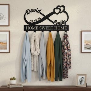 May include: Black metal wall-mounted coat rack with an infinity symbol design, the names "David" and "Emily", and the date "07.15.24". The rack has hooks holding various sweaters and a scarf. The text "HOME SWEET HOME" is below the infinity symbol.