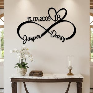 Pode incluir: Um letreiro de parede em metal preto com um símbolo de infinito entrelaçado com um formato de coração. O design inclui os nomes Jasper e Daisy em letra cursiva, a data 15.07.2005 e o número 18, integrados na estrutura de linha preta.