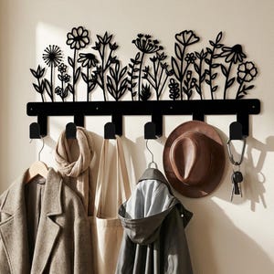 Könnte beinhalten: Eine schwarze Wandgarderobe aus Metall mit einer dekorativen Silhouette aus verschiedenen Wildblumen und Stängeln an der Oberseite. Die Leiste verfügt über sechs Haken, an denen ein Mantel, ein Schal, eine Tasche, ein Hut und ein Schlüsselbund hängen.