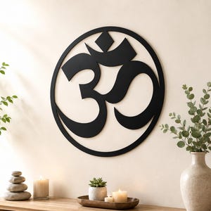 Könnte beinhalten: Ein kreisförmiges Wandkunstobjekt aus schwarzem Metall mit dem Om-Symbol. Das Design besteht aus einem massiven Außenring, der das stilisierte Sanskrit-Zeichen umschließt, welches eine Mondsichelform und einen rautenförmigen Punkt oben mittig enthält.