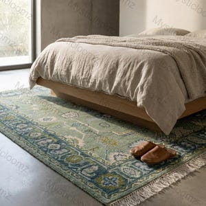 May include: A rectangular, green and blue patterned rug with a fringed edge is placed on a concrete floor in front of a bed. The rug has a detailed border design and is partially under a wooden bed frame. Brown leather slippers are on the rug.