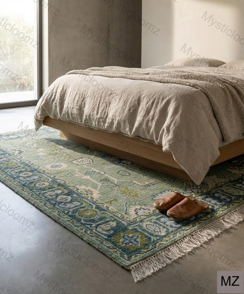 May include: A rectangular, green and blue patterned rug with a fringed edge is placed on a concrete floor in front of a bed. The rug has a detailed border design and is partially under a wooden bed frame. Brown leather slippers are on the rug.