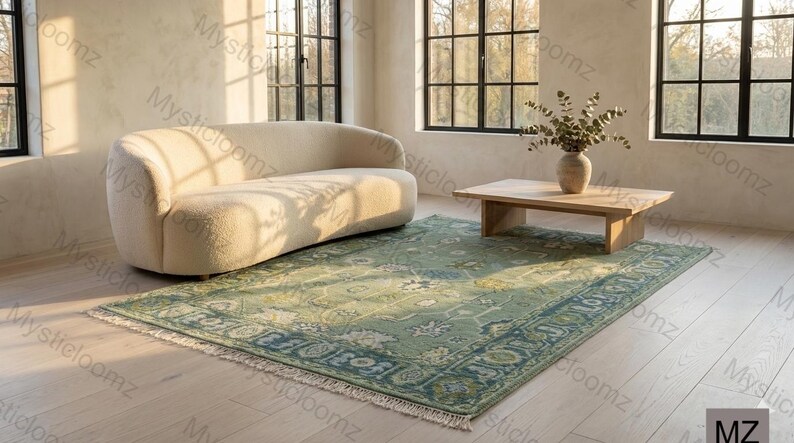 May include: A light green and blue patterned rug with a cream-colored fringe sits on a light wood floor. A curved cream-colored sofa and a wooden coffee table are in the background. The room has large windows.