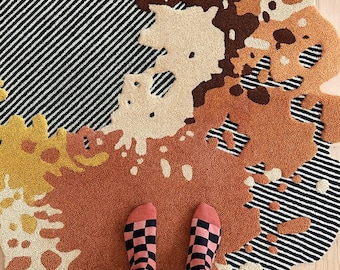 May include: A colorful, abstract rug with a unique, organic shape. The rug features a mix of patterns, including black and white stripes, spots, and solid colors in shades of brown, beige, and orange. A person's feet wearing checkered socks and striped pants are visible.