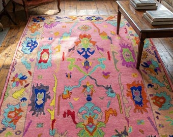 May include: A vibrant pink area rug with a detailed, colorful pattern. The rug features a central pink field with a border of beige and a complex design in shades of blue, orange, green, and purple. The rug is placed on a wooden floor.