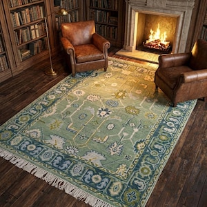 May include: A rectangular, light green rug with a detailed floral pattern and a dark blue border. The rug is in a room with two brown leather armchairs, a fireplace, and bookshelves filled with books.