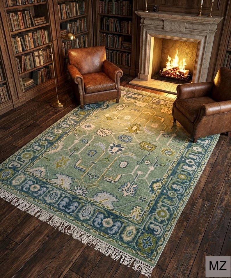 May include: A rectangular, light green rug with a detailed floral pattern and a dark blue border. The rug is in a room with two brown leather armchairs, a fireplace, and bookshelves filled with books.