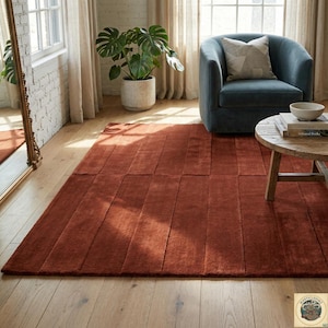 Pode incluir: Um tapete retangular cor de ferrugem com um padrão vertical texturizado. O tapete é colocado sobre um chão de madeira numa sala bem iluminada, com uma poltrona azul e uma mesa redonda de madeira por perto. As dimensões do tapete não são visíveis.