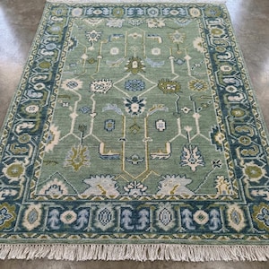 May include: A rectangular, sage green rug with an intricate geometric pattern. The rug features a dark teal border with repeating floral and geometric designs. White fringe trims the edges. The rug's central design includes floral and geometric shapes in shades of blue, green, and cream.