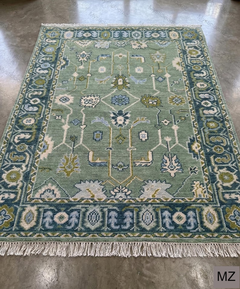 May include: A rectangular, sage green rug with an intricate geometric pattern. The rug features a dark teal border with repeating floral and geometric designs. White fringe trims the edges. The rug's central design includes floral and geometric shapes in shades of blue, green, and cream.