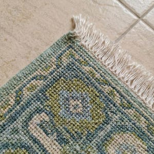 May include: Close-up of a section of a rug with a geometric pattern in shades of blue, green, and cream. The rug has a textured appearance and a fringe edge. The corner of the rug is visible.