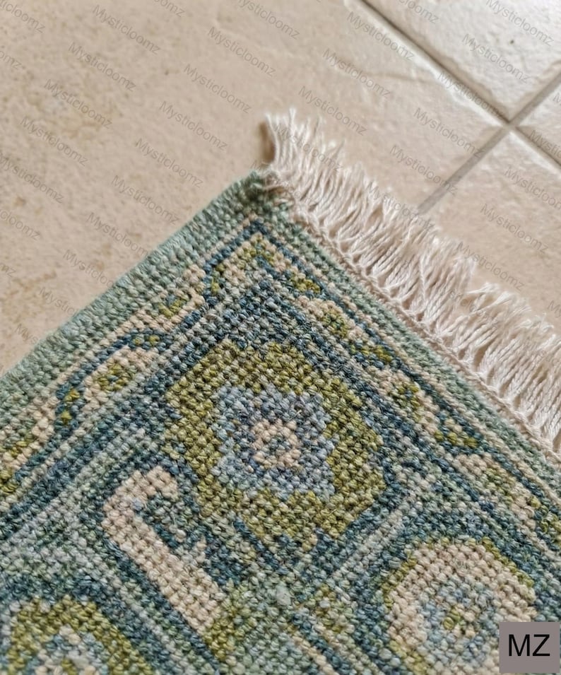 May include: Close-up of a section of a rug with a geometric pattern in shades of blue, green, and cream. The rug has a textured appearance and a fringe edge. The corner of the rug is visible.