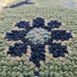 May include: Close-up of a hand-tufted rug with a floral design. The rug features a central flower in shades of blue and light blue, surrounded by a green and off-white background. The texture is dense and the craftsmanship is evident.