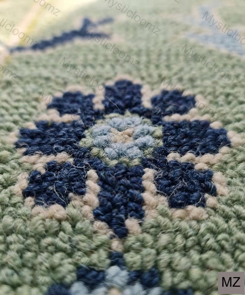 May include: Close-up of a hand-tufted rug with a floral design. The rug features a central flower in shades of blue and light blue, surrounded by a green and off-white background. The texture is dense and the craftsmanship is evident.