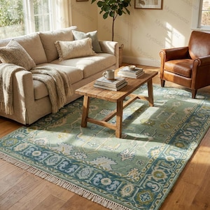 May include: A light green and blue patterned area rug with a cream fringe is placed on a hardwood floor. The rug features a central floral design and a border with geometric patterns. A wooden coffee table, sofa, and leather armchair are in the background.