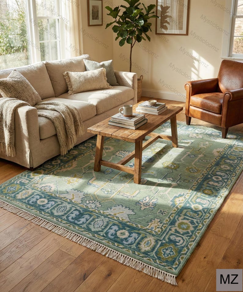 May include: A light green and blue patterned area rug with a cream fringe is placed on a hardwood floor. The rug features a central floral design and a border with geometric patterns. A wooden coffee table, sofa, and leather armchair are in the background.