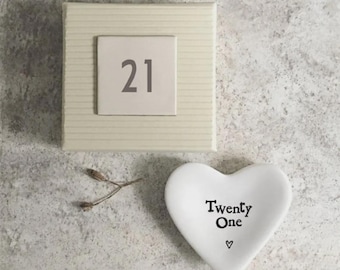 May include: A square, cream-colored box with the number "21" on a smaller square. A white heart-shaped dish with the words "Twenty One" and a small heart symbol is also present. A sprig of dried plant material is next to the dish.