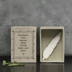 Puede incluir: Una caja de cerillas decorativa con el texto "Feathers will appear when loved ones are near" y una pluma blanca en su interior. La caja de cerillas está abierta, revelando la pluma. Un pequeño acento floral verde está en primer plano.