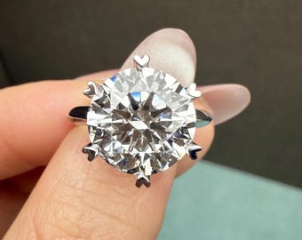 May include: A silver-toned ring featuring a large, round, brilliant-cut clear gemstone as the centrepiece. The stone is secured by six prongs, each uniquely shaped like a small heart at the tip, set against a polished metallic band.