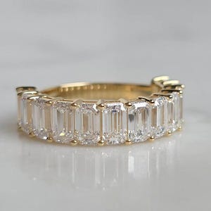 May include: A gold band ring featuring a continuous row of clear, rectangular emerald-cut stones set in a prong setting. The ring displays a polished yellow gold metal finish and a series of faceted, transparent stones arranged side by side.