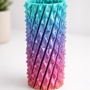 May include: A tall, cylindrical vase with a textured surface and a gradient color scheme. The vase transitions from turquoise at the top to purple and then to pink at the base. The surface has a repeating pattern of triangular spikes.