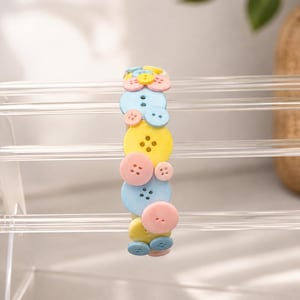 May include: A headband decorated with a variety of round, four-hole buttons in pastel shades of light blue, soft pink, and bright yellow. The buttons are arranged in a dense, overlapping pattern along the entire length of the band.