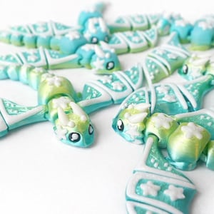 May include: A collection of handmade, articulated dragon figurines in shades of turquoise, green, and white. Each dragon features intricate details, including small horns, eyes, and star-shaped accents. The dragons are arranged on a white surface.