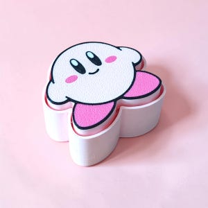 Könnte beinhalten: Ein weiß-rosa Kirby-förmiger Behälter mit einem lächelnden Gesicht und rosa Wangen. Der Behälter hat eine weiße Oberseite mit schwarzer Umrandung und rosa Akzenten. Der Hintergrund ist einfarbig rosa.