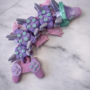 May include: A colourful sculpture in the shape of a video game controller and a dragon. The sculpture is made of purple, pink, silver, and teal elements. The controller buttons are teal. The dragon-like form has multiple segments.