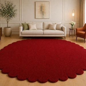 May include: A large, round, red rug with a scalloped edge dominates the room. The rug is placed on a hardwood floor in front of a cream-colored sofa and an orange armchair. A potted plant and artwork are also visible.