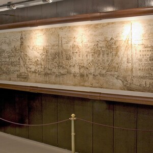 May include: A large, framed artwork showing a detailed harbour scene with sailboats and buildings. The artwork is displayed on a dark wood-panelled wall, with two people standing in front of it. Additional framed images are visible to the right.