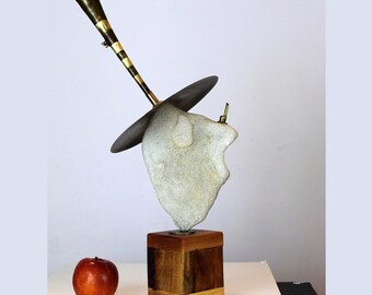 Könnte beinhalten: Abstrakte Skulptur mit einem Steinkopf mit Hut, einem Messinghorn und einem Holzsockel. Das Horn hat schwarz-goldene Streifen. Ein roter Apfel liegt vor der Skulptur auf der weißen Oberfläche.