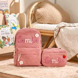 May include: A pink corduroy backpack and matching lunch bag set. The backpack and lunch bag have the name "Camila" embroidered on them in a cursive font. The backpack has a flower applique and the lunch bag has a strawberry applique.