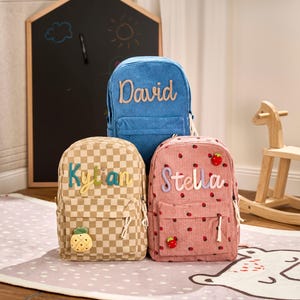 May include: Three personalized backpacks in a child's room. One is blue corduroy with "David" stitched on it. Another is checkered with "Kylan" and a pineapple. The third is pink with strawberries and "Stella".