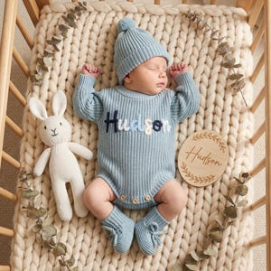 May include: A sleeping baby wearing a light blue knit romper, hat, and booties, with the name "Hudson" embroidered on the chest. A white stuffed bunny and a wooden name plaque are also in the crib. The baby is lying on a cream-colored knitted blanket.
