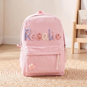 May include: A light pink corduroy backpack featuring the name Rosalie in raised, multi-coloured yarn lettering across the front. The bag includes a front zippered pocket adorned with a small, embroidered pink flower and a white cord zipper pull.