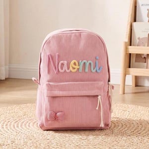 May include: A pink corduroy backpack featuring the name Naomi in colourful, raised, rounded letters across the top. The front pocket includes a small red and white checkered bow detail and a white cord zipper pull on the side of the bag.